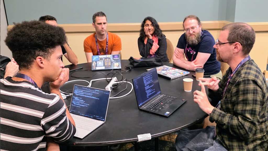 Attendees gather around a round table during the pyOpenSci generative AI open space at PyCon US 2026, laptops open as one participant gestures while leading a small-group discussion.