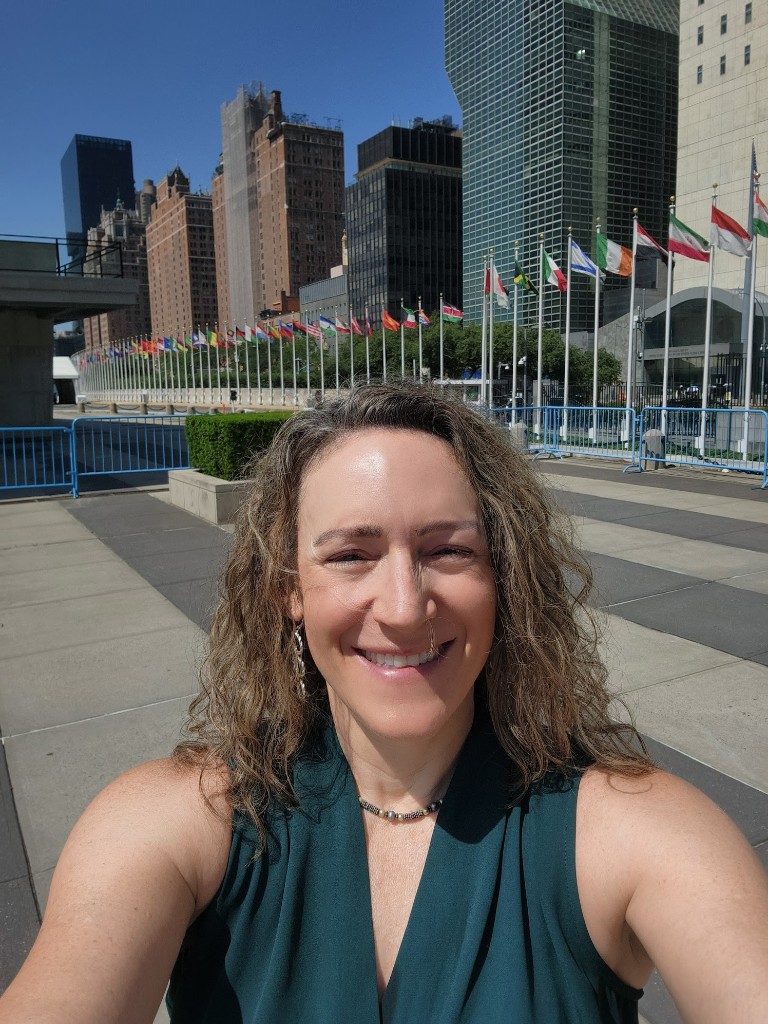 Leah Wasser smiling in front of the United Nations Headquarters in New York City, with international flags flying behind her.
