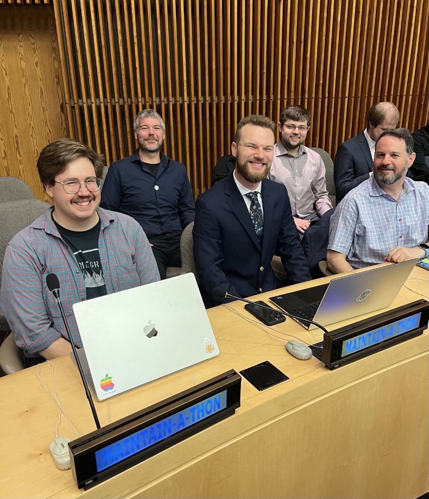 Open source maintainers seated at a conference desk with laptops during the Maintain-a-thon at UN Open Source Week. Blue desk signs read Maintain-a-thon.