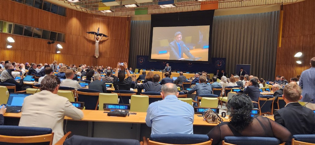 A large assembly hall at the United Nations filled with attendees during an OSPO for Good session. A speaker presents at the front while blue desk signs read OSPO for Good.