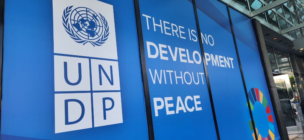 A blue United Nations Development Programme sign at UN Headquarters reading There is no development without peace, with the UN and Sustainable Development Goals logos visible.